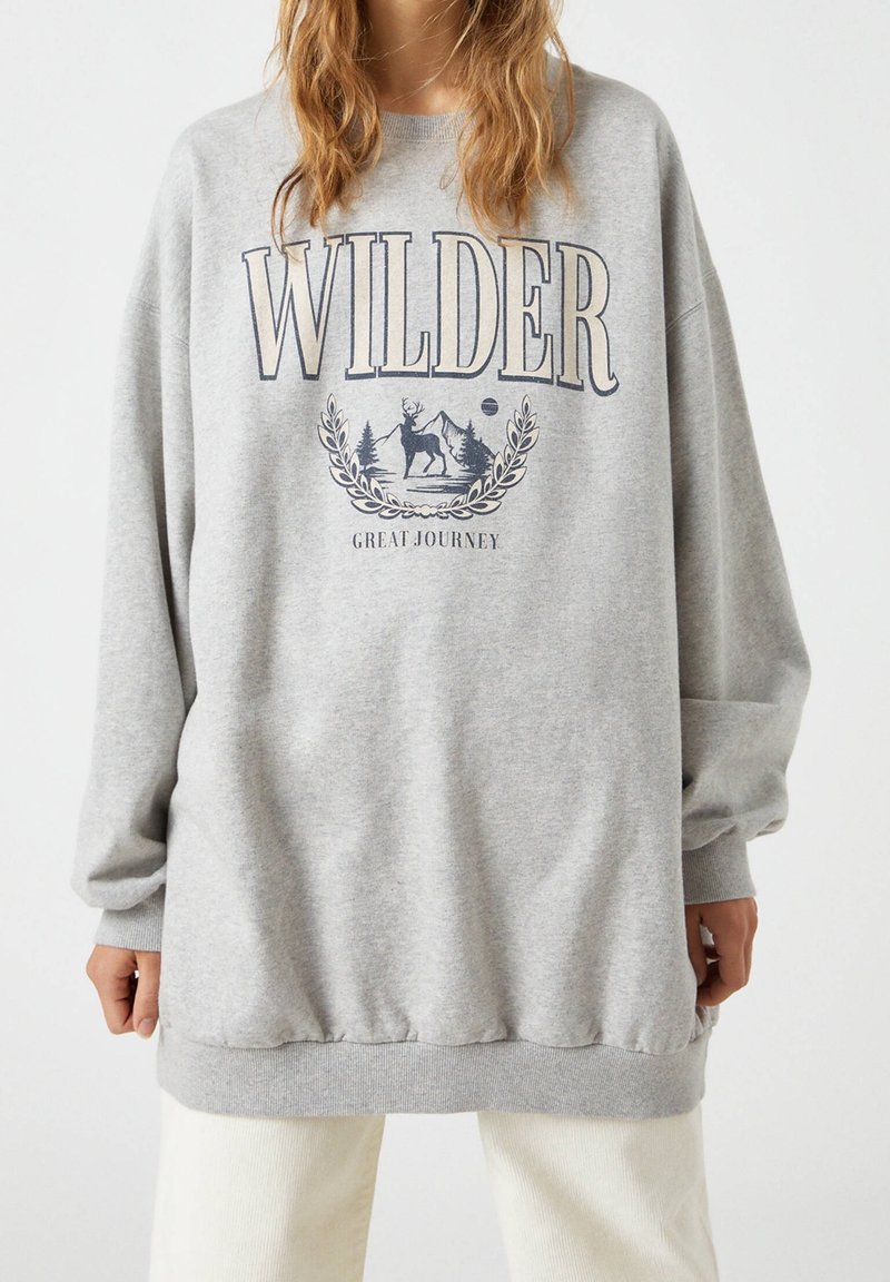Personne portant un sweat-shirt gris oversize avec le texte "WILDER" et une illustration de cerf, montagnes et arbres au-dessus du texte "GREAT JOURNEY".
