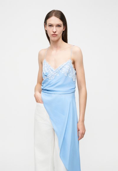 Γυναίκα που φοράει ανοιχτό μπλε καμησόλ με δαντέλα και ασύμμετρο τελείωμα, συνδυασμένο με άσπρο παντελόνι φαρδιάς γραμμής, στέκεται μπροστά σε λευκό φόντο.