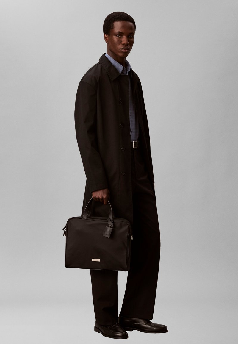 Άνδρας που φοράει μαύρο παλτό, σκούρο παντελόνι, μπλε πουκάμισο, κρατώντας μαύρη τσάντα εργασίας, στέκεται μπροστά σε γκρι φόντο.