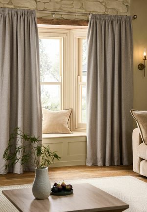 Beige vinduesgardiner delvist, der dækker en karnap med en beige pude på sædet, ved siden af en beige sofa og et bord i træ med en vase.