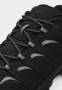 Μαύρο αθλητικό παπούτσι με χοντρές, στρογγυλές κορδόνια με σχέδιο, περασμένα μέσα από υφασμάτινες θηλιές και σουέντ λουράκια πάνω από διαπνέοντα πλέγματα.