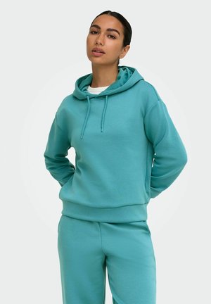 Jeune femme les mains derrière le dos, portant un sweat à capuche bleu sarcelle et un pantalon assorti, face à un fond uni.