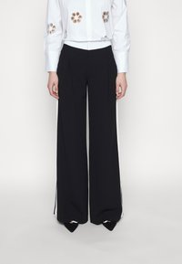 Svarta vida byxor med en slät yta, prydda med en vit sidostrip. Designen inkluderar en liten spricka vid fållen för bättre rörelse.