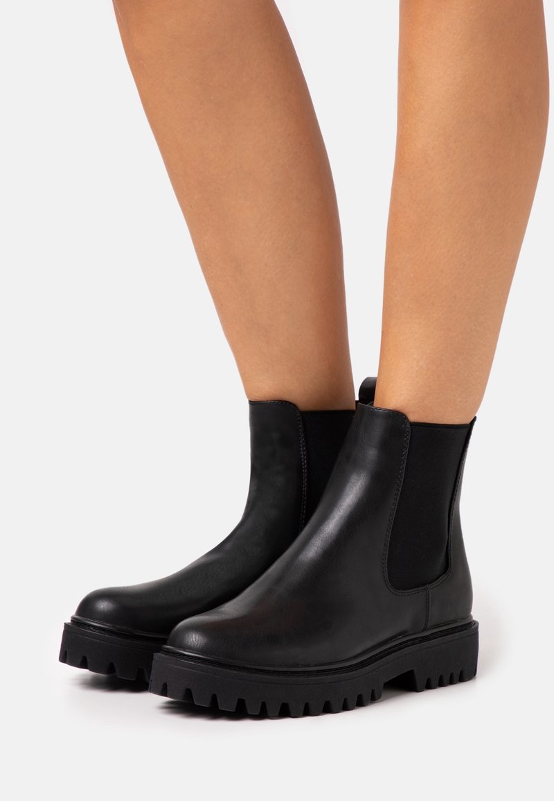 Bottines en cuir noir avec semelles épaisses à crampons portées sur des jambes nues sur un fond clair uni.