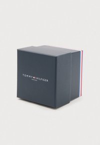 Ciemnoniebieskie pudełko na zegarek Tommy Hilfiger z logo i akcentami w czerwono, białym i niebieskim kolorze na białym tle.