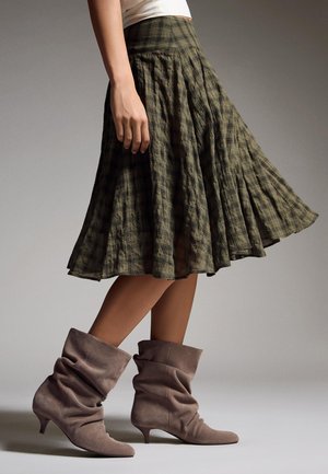 Kvinde iført et knælængde olivengrønt ternet plisseret skørt og løstsiddende taupe ruskindsankelstøvler med lave hæle.