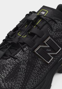 Gros plan d'une chaussure de sport noire avec un motif texturé, des lacets élastiques avec un mécanisme de verrouillage et un grand logo en relief sur le côté.