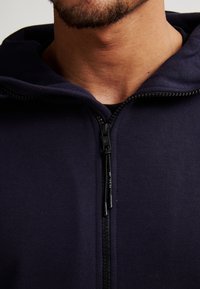 Sudadera con cremallera en color azul marino, con una textura suave, que cuenta con una cremallera frontal y una capucha ajustable. La vista en primer plano destaca el detalle del cuello y la cremallera.