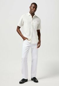 Chemise en lin écrue à manches courtes avec fermeture à boutons et poche poitrine, associée à un pantalon blanc et des mocassins noirs.