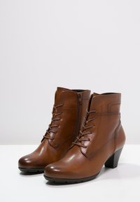 Botas de tobillo de cuero marrón con tacón apilado, que presentan un diseño de cordones en la parte delantera y una cremallera lateral para facilitar su uso. Textura suave.