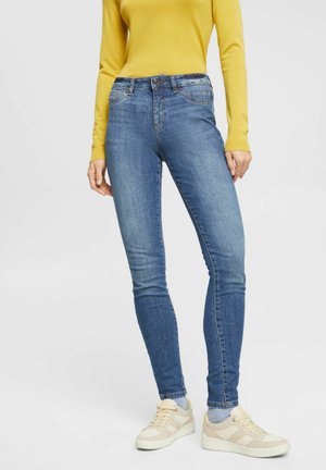 Osoba ubrana w dopasowane niebieskie dżinsy, żółtą bluzkę z długim rękawem i beżowe trampki, stojąca z rękami swobodnie opartymi wzdłuż ciała.