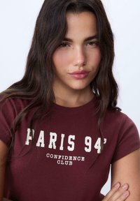 Těsně přiléhající vínové tričko s krátkými rukávy, na přední straně s bílým grafickým potiskem "PARIS 94' CONFIDENCE CLUB" a hladkou texturou.