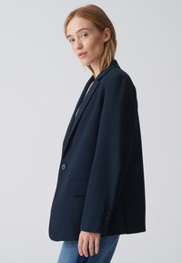 Blazer marine, design ajusté, fermeture à bouton unique, deux poches avant, manches longues avec accents de boutons, texture de tissu lisse.