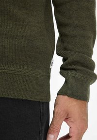 Maglione verde lavorato a maglia con polsini e orlo a costine, caratterizzato da una finitura testurizzata. Include una piccola etichetta del marchio vicino alla cucitura.