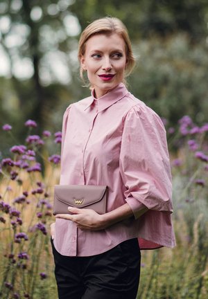 Pink, collared blouse with puff sleeves, paired with a taupe clutch featuring a gold accent. Set against a garden backdrop.