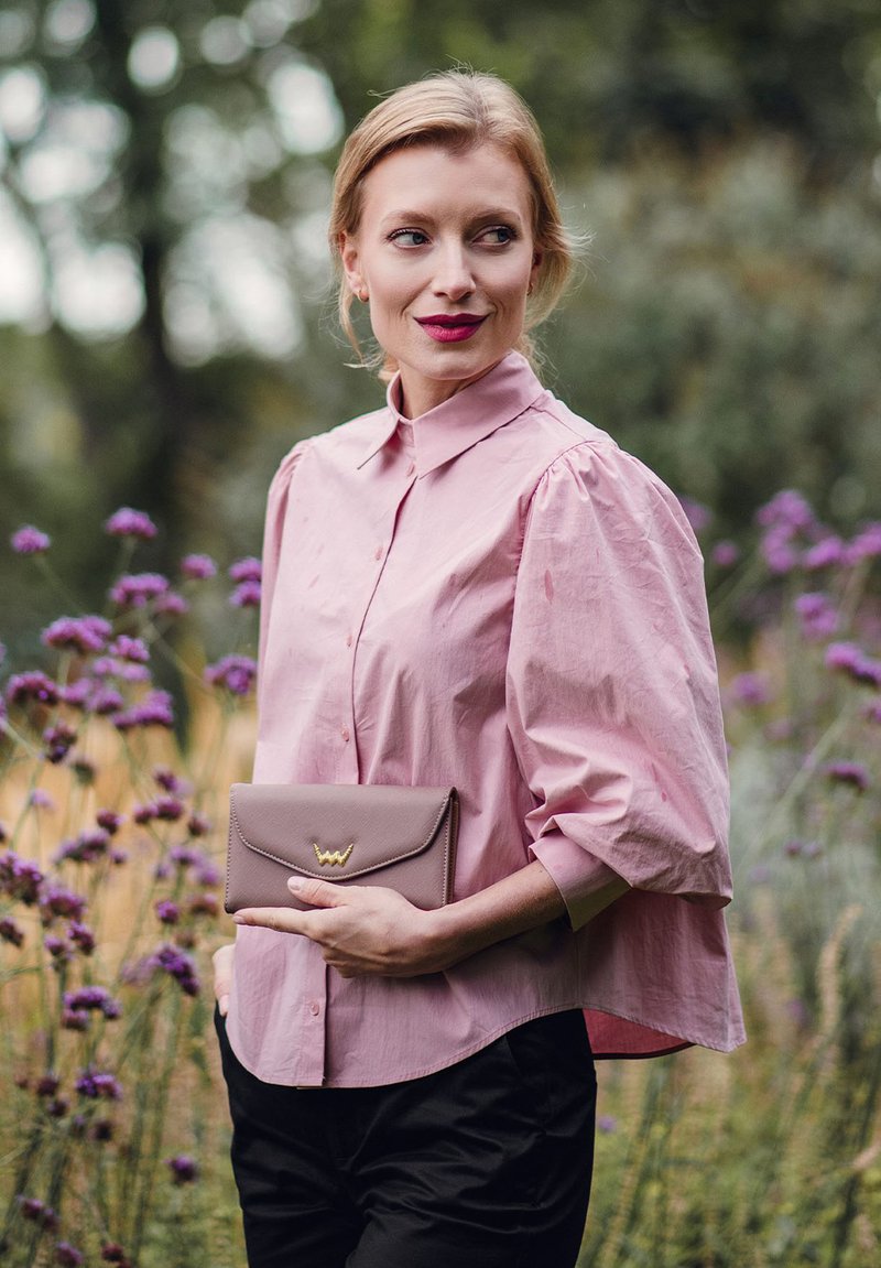 Pink, collared blouse with puff sleeves, paired with a taupe clutch featuring a gold accent. Set against a garden backdrop.