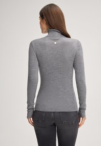 Maglione a collo alto grigio a costine con maniche lunghe, caratterizzato da un piccolo dettaglio a bottone metallico sul retro, abbinato a jeans scuri.