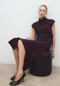 Mujer sentada en un taburete redondo negro, vestida con un vestido sin mangas de cuello alto de color púrpura oscuro y tacones altos negros, frente a una pared blanca.
