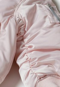 Manche de bomber en satin rose clair avec texture froncée, présentant une fermeture éclair argentée, un poignet côtelé et une finition lisse.
