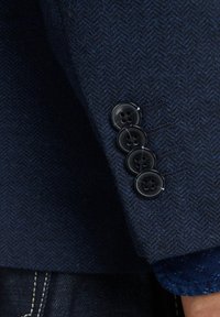 Manica di un blazer strutturato blu navy con quattro bottoni neri opachi e un motivo a spina di pesce sul tessuto.