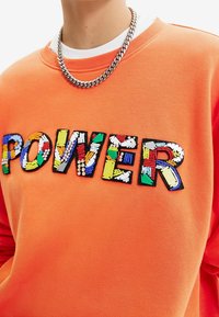 Oranje sweatshirt met het woord "POWER" in meerkleurige textuurletters, tegen een achtergrond van gladde stof. Zilveren ketting.