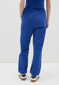 Blauwe katoenen joggers met een elastische tailleband en taps toelopende benen, voorzien van een gladde textuur en subtiele glans. G gedragen met witte en marineblauwe sneakers.