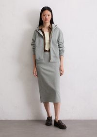 Haut à capuche zippé gris porté sur un débardeur beige, associé à une jupe crayon grise. Le mannequin porte des chaussures à enfiler sombres ; fond uni.