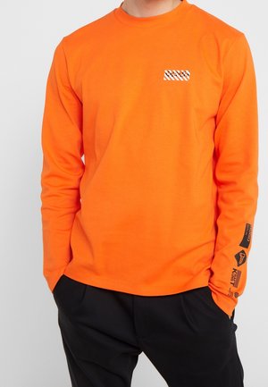 Person trägt ein leuchtend oranges Langarmshirt mit kleinem weiß-schwarz gestreiftem Logo auf der Brust und schwarzen Druckmotiven am unteren rechten Ärmel.