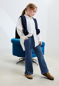 Biela košeľa na zapínanie s tmavomodrým svetrom prehodeným cez plecia. Slim-fit tmavomodré rifle s bočnými rázporkami, hnedé mokasíny na slip-on.