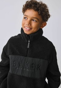 Zwarte fleece pullover met hoge kraag en halve rits. Heeft een glad paneel met "BAKER BY TED BAKER" in het zwart geperst.