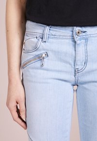 Jeans de mezclilla azul claro con un bolsillo con cremallera en la parte delantera, diseño de cinco bolsillos y detalles de costura sutiles. Textura suave, forma ajustada.