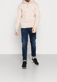 Uomo che indossa una felpa beige con cappuccio, jeans blu scuri arrotolati, calzini bianchi e sneaker nere, in piedi su un pavimento grigio contro un muro bianco.