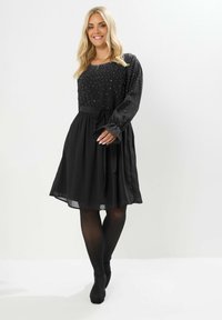 Musta mekko pitkillä hihoilla, jossa on helmitoppi, vyötärö kiristetty nauhalla ja liehuva hame. Silkkinen kangas, jossa on läpikuultava kerros.