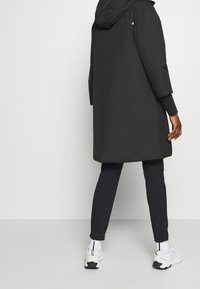 Manteau imperméable noir avec capuche, coupe mi-longue et manches longues, associé à un legging noir et des baskets blanches texturées.