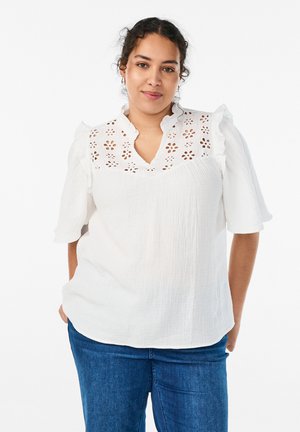 Ženska s temnimi kodrastimi lasmi nosi belo bluzo z cvetličnimi izrezi in nagubaniimi rokavi, v kombinaciji z modrimi kavbojkami, stoji na belem ozadju.