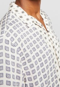 Lyhythihainen paita kevyestä kankaasta, jossa on valkoinen tausta ja sinisiä geometrisia kuvioita, varustettuna nappikauluksella ja rentoa istuvuutta.