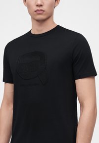Νέος άνδρας που φοράει μαύρο t-shirt με υφασμένη γραφική παράσταση ενός στυλιζαρισμένου προσώπου με γυαλιά ηλίου και ουρά αλόγου στο στήθος.