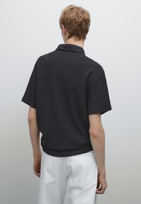 Černý polo tričko s krátkým rukávem a texturovanou tkaninou, vybavené límcem. Tričko je zkombinováno s bílými kalhotami, což vytváří uvolněný vzhled.