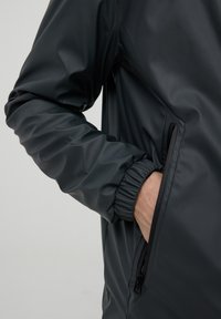 Giacca grigia scura idrorepellente con polsini elastici, tasca laterale con zip e texture liscia. Presenta un design minimalista e una silhouette snella.