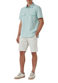Kurzärmliges, hellmintgrünes Button-Up-Hemd mit zwei Brusttaschen, kombiniert mit weißen Shorts und beigen Sneakers mit braunen Akzenten.