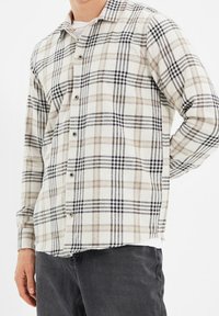 Camicia a quadri in beige, nero e crema, realizzata in tessuto testurizzato. Presenta chiusura con bottoni, maniche lunghe e un colletto con vestibilità rilassata.