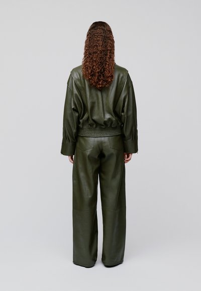 Persoană cu păr creț și castaniu, purtând o jachetă din piele verde închis și pantaloni cu picioare largi, stând cu fața spre spate pe un fundal simplu.