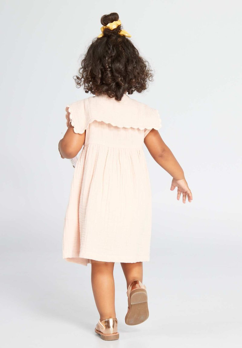 Kleinkind mit lockigem Haar, das ein hellrosa Kleid und glänzende roségoldene Schuhe trägt und auf einem einfarbigen hellen Hintergrund davon geht.