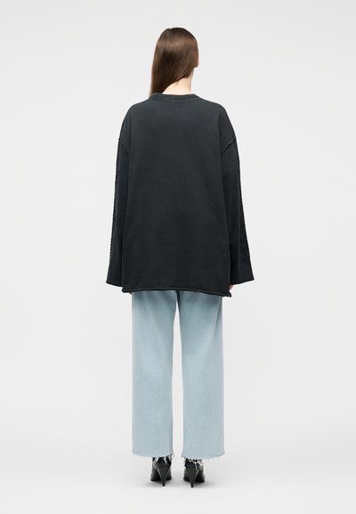 Άτομο με μακριά καστανά μαλλιά που φοράει ένα υπερμεγέθες μαύρο φούτερ, ανοιχτόχρωμο τζιν παντελόνι με ευρεία μπατζάκια και μαύρα παπούτσια με μυτερή φτέρνα, σε θέα από πίσω.