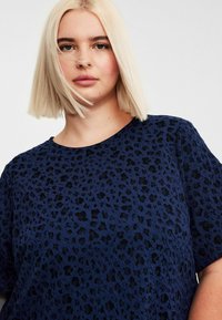 Blonde Frau mit glattem Haar, die ein blaues und schwarzes gemustertes, strukturiertes Kurzarmoberteil trägt und frontal vor einem schlichten Hintergrund steht.
