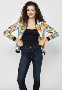 Giacca bomber floreale con motivi gialli e blu, polsini e orlo in maglia nera, indossata sopra una maglietta nera, abbinata a jeans di denim scuro.