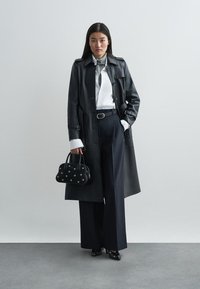 Black leather trench coat over a white shirt, paired with high-waisted navy wide-leg pants. Accessorized with a small black bag featuring silver studs.