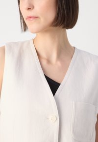 Lichtbeige mouwloze vest met een v-hals, enkele knoopsluiting, een opgestikte zak aan de voorkant en een gestructureerde stof.
