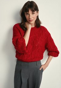 Czerwony sweter w splot warkoczowy z okrągłym dekoltem i luźnymi rękawami, zestawiony z szarymi spodniami w prążki. Faktura jest gruba i miękka.