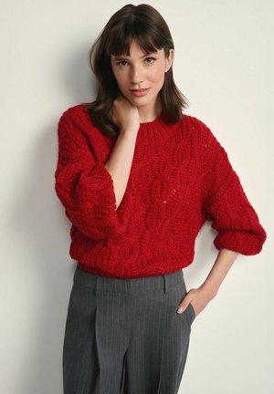 Czerwony sweter w splot warkoczowy z okrągłym dekoltem i luźnymi rękawami, zestawiony z szarymi spodniami w prążki. Faktura jest gruba i miękka.